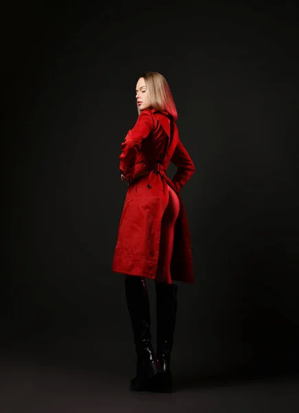 Захоплена розкішна молода блондинка в шкіряних високих чоботях і червоне пальто з оголеною дупою стоїть на камеру, виходячи — стокове фото