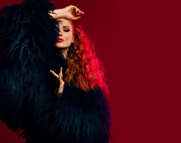 Underbar rik ung kvinna med rött lockigt hår bär lyx päls poserar med slutna ögon, arm i huvud över rött — Stockfoto