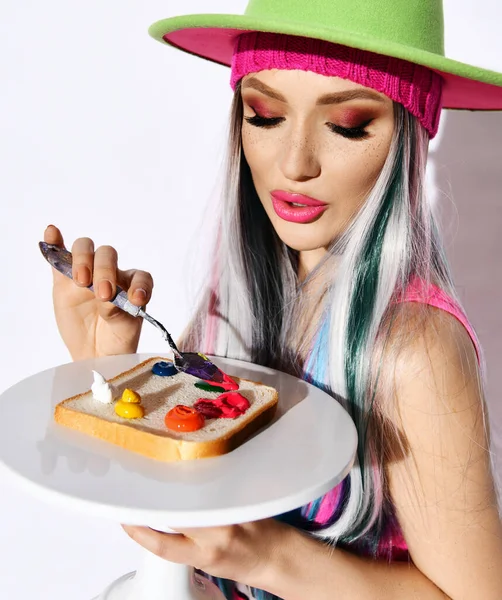 Fashionable woman modern artist in green hat holds tray with bread, smears oil paints on it with spatula, going to eat — Stock Photo, Image