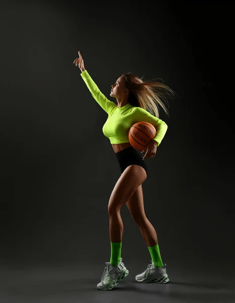 Sporty woman in sportswear, top, shorts, sneakers stands holding basketball ball in hand and holding the other hand up — Stock Photo, Image