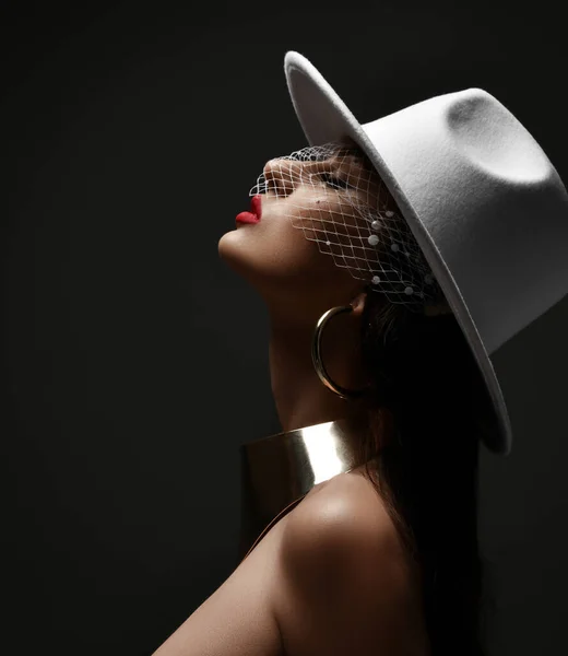 Profile of rich woman in white hat with veiling and massive metal collar and earrings holding head up with eyes closed