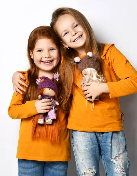Happy meninas pequenas irmãs ou amigos em terracota amarelo confortáveis mangas compridas de pé abraçando e segurando bonecas de brinquedo — Fotografia de Stock