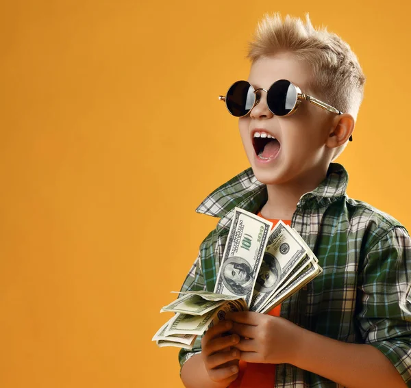 Blonde rich kid boy in round sunglasses and checkered plaid shirt stands with fan of dollar cash in hands and screams — стоковое фото