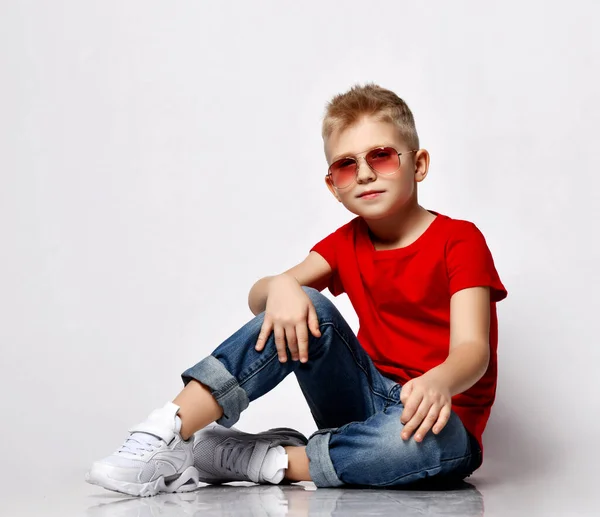 Cool blonde kid boy leader in red t-shirt, blue jeans, white sneakers and sunglasses sits on floor in relaxed pose — Zdjęcie stockowe