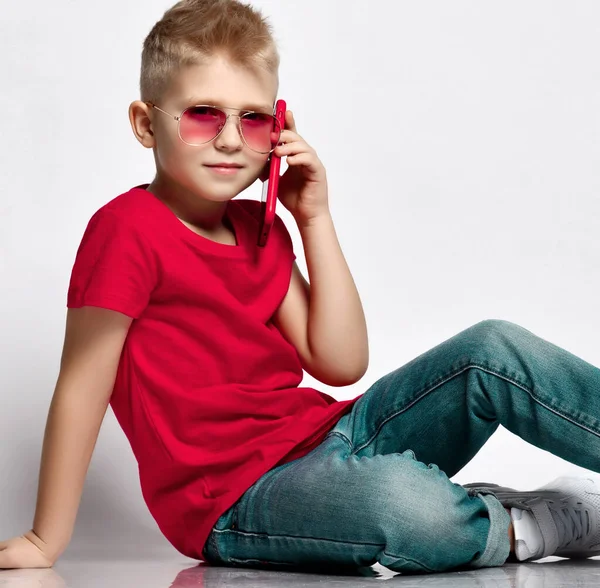 Stylish kid boy child in red t-shirt, jeans, sneakers and sunglasses sitting on floor talking on smartphone — Zdjęcie stockowe