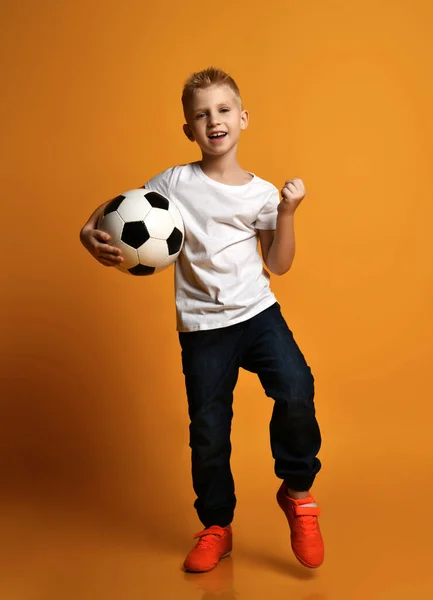 Blond positive boy child in white t-shirt and jeans standing with soccer ball in hand and showing win sign with hand