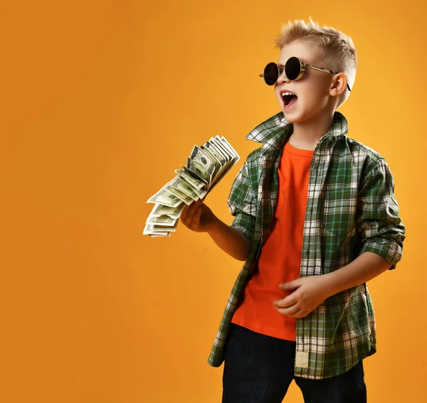 Blond smiling boy child in checkered shirt and sunglasses standing holding heap of money cash dollars in hands having fun —  Fotos de Stock