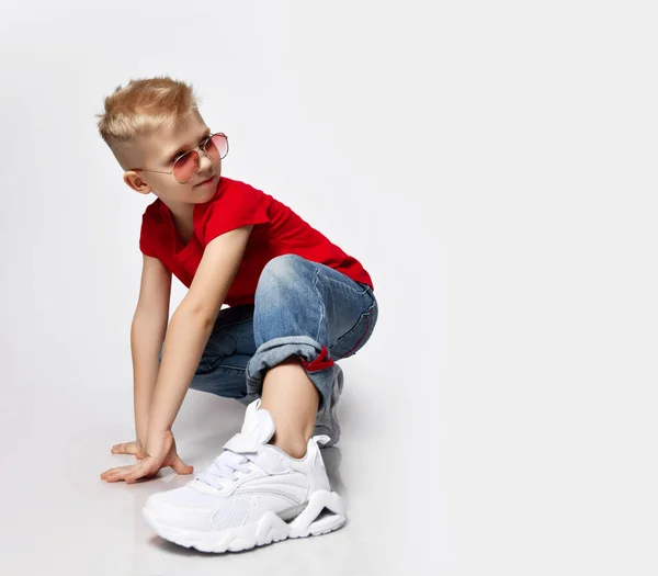 Active, frolic blonde kid boy in red t-shirt, blue jeans, white sneakers and sunglasses is in ready to run position — Stock fotografie