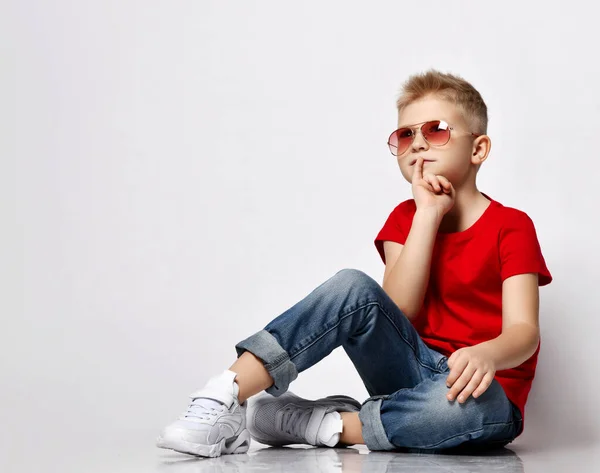 Elegante niño rubio líder en camiseta roja, vaqueros azules, zapatillas blancas y gafas de sol se sienta en el suelo pensativo —  Fotos de Stock