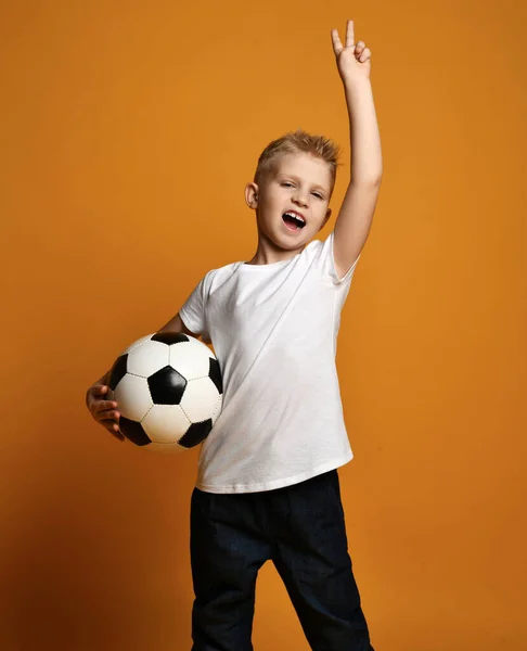 Gritando em voz alta menino loiro em branco t-shirt stands segurando bola de futebol na mão e gestos V sinal de vitória — Fotografia de Stock