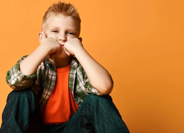 Calma adolescente colegial, chico normal en camisa a cuadros, naranja camiseta y jeans se sienta sosteniendo puños en sus mejillas —  Fotos de Stock