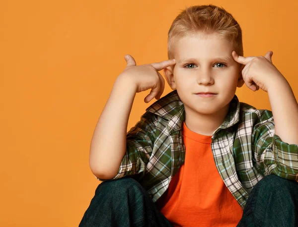 Colegial adolescente en camisa a cuadros, naranja camiseta y vaqueros se sienta sosteniendo los dedos en la cabeza, haciendo un gesto piénsalo —  Fotos de Stock