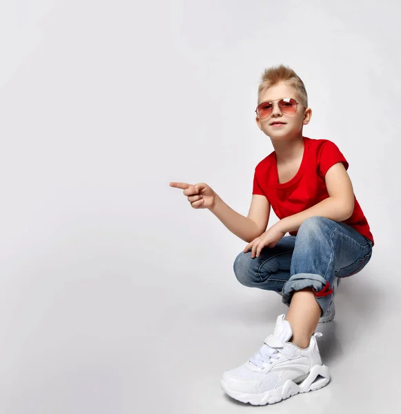 Enfant blond frolic en t-shirt rouge, jean bleu, baskets et lunettes de soleil assis accroupi pointant du doigt l'espace de copie — Photo