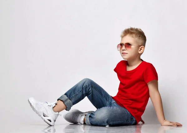 Pravidelný blonďatý chlapec v červeném tričku, modrých džínách, bílých teniskách a brýlích sedí na podlaze a opírá se o ruce — Stock fotografie