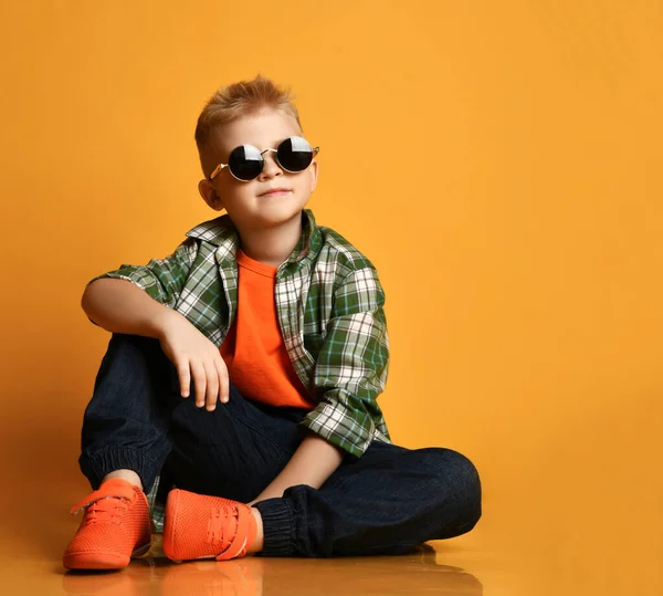 Spokojny miły dzieciak, nastolatek w okrągłych okularach przeciwsłonecznych, w kratkę, koszulce i dżinsach siedzi na podłodze patrząc w górę — Zdjęcie stockowe