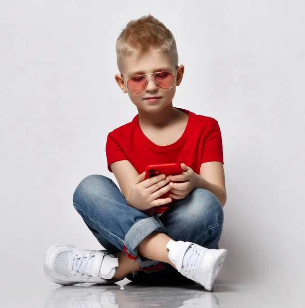 Stylish kid boy child in red t-shirt, jeans, sneakers and sunglasses sitting on floor with legs crossed looking at smartphone — Stock Photo, Image