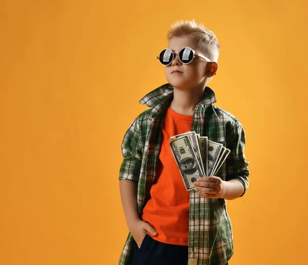 Tranquilo adolescente rico de confianza en sí mismo en gafas de sol redondas, camisa a cuadros y pantalones vaqueros se mantiene ventilador de efectivo —  Fotos de Stock