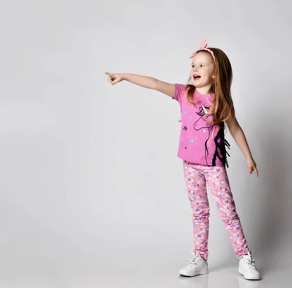 Happy screaming blonde kid girl in summer clothing colorful pants and t-shirt stands pointing with finger at copy space — Stock Photo, Image