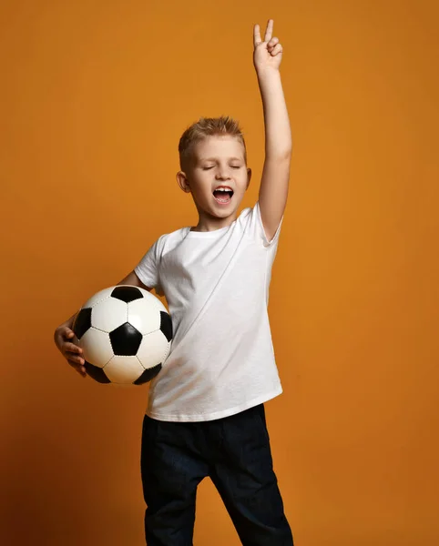 Menino feliz em branco t-shirt fica segurando bola de futebol na mão e gestos V sinal de vitória, celebra vitória — Fotografia de Stock