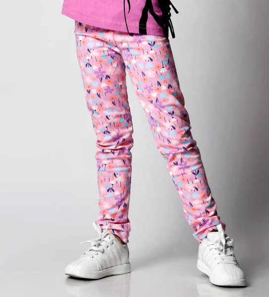 Zbliżenie dziewczynki w kolorowych różowych spodniach z kwiatowym wzorem, trampki stojące nad jasnym tłem — Zdjęcie stockowe