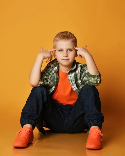 Chytrý školák teenager v kostkované košili, oranžovém tričku a džínách sedí na podlaze a drží prsty u hlavy a ukazuje — Stock fotografie