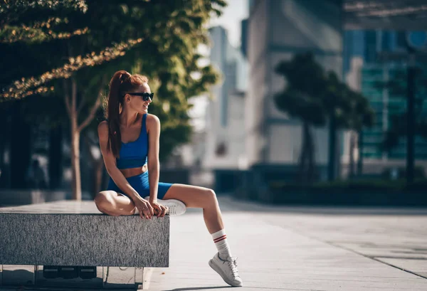 Χαμογελώντας ευτυχισμένη αθλητής γυναίκα στο δρομέα αθλητικών ειδών κάθεται στον πάγκο στην πόλη μετά από ασκήσεις προπόνηση και κοιτάζει στην άκρη στο Ντουμπάι — Φωτογραφία Αρχείου