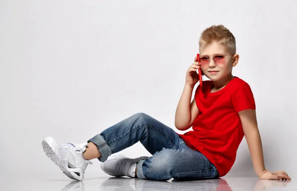 Cool niño rubio en camiseta roja, vaqueros azules, zapatillas blancas y gafas de sol se sienta en el suelo hablando por teléfono celular —  Fotos de Stock