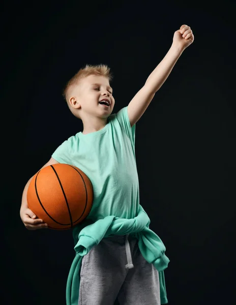 Cheerful happy kid boy in t-shirt, pants and hoodie around waist stands holding basketball ball and arm up outstretched — Stock Photo, Image