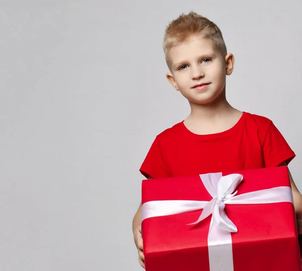 Portrait de garçon souriant en t-shirt rouge tenant une grande boîte cadeau avec ruban dans les mains, le portant, prêt pour l'anniversaire — Photo