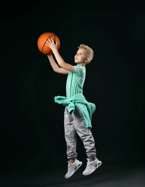 Niño rubio en camiseta verde, pantalones, zapatillas y sudadera con capucha alrededor de la cintura salta lanzando la pelota de baloncesto —  Fotos de Stock