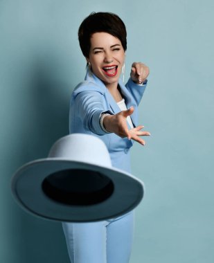 Çekici, kısa saçlı, esmer, mavi iş elbiseli şık takım elbiseli kadın kameraya şapka fırlatıyor.