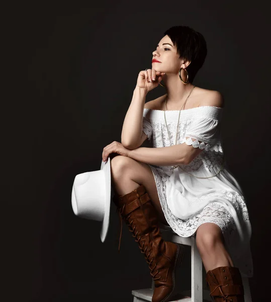 Ung positiv ganska kort hår brunett kvinna i vit elegant klänning stövlar och hatt sitter på pall tittar åt sidan — Stockfoto