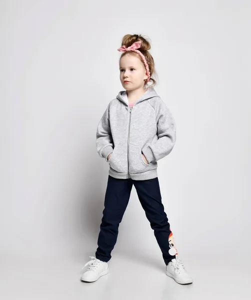 Naughty, daring, self-confident kid girl in gray hoodie with zipper standing with hands in pockets and looking aside — Stock Photo, Image
