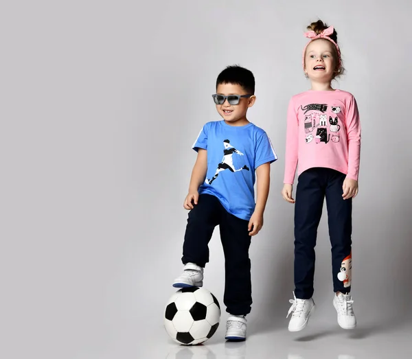 Happy active kids in summer clothes, Asian boy stands with soccer ball while Caucasian blonde girl jumumps near him — Fotografia de Stock