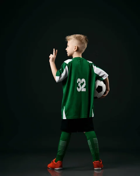 Estudante da escola de futebol, adolescente de uniforme listrado branco vermelho fica de volta à câmera segurando bola e gesticulando V sinal — Fotografia de Stock