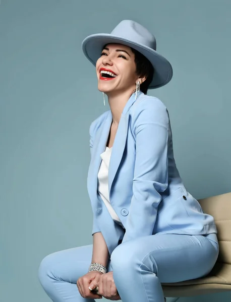 Cheerful brunette woman in blue tight business suit and wide-brimmed hat sits on bar stool happy laughing — Stock Photo, Image