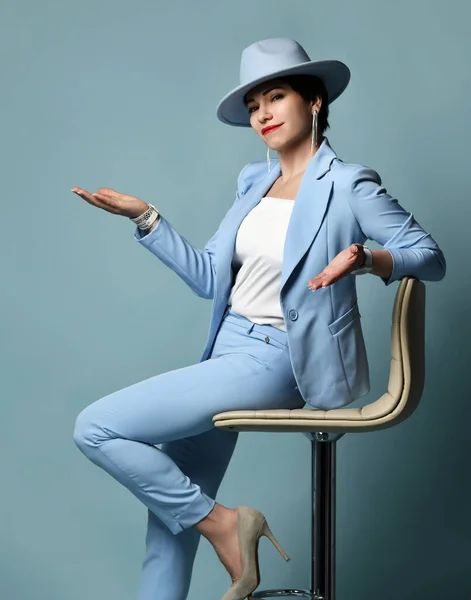 Sorrindo mulher morena de cabelos curtos em terno de negócios azul e chapéu senta-se no banquinho de mãos dadas com a palma aberta — Fotografia de Stock