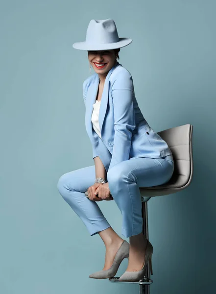 Cheerful brunette woman in blue business suit, covering eyes hat and high-heeled shoes sits on bar stool. Full-growth — Stock Photo, Image
