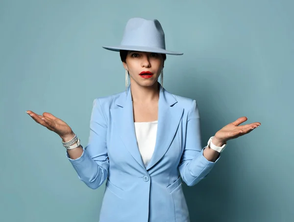 Frustrated short haired brunette businesswoman in blue official suit and hat stands holding hands up, palms up — Stock Photo, Image