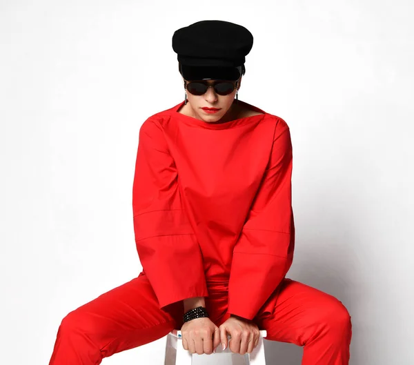 Стильные короткие волосы брюнетка женщина в высокой моды случайный красный брючный костюм, солнцезащитные очки и черная кепка сидит с раздвинутыми ногами — стоковое фото