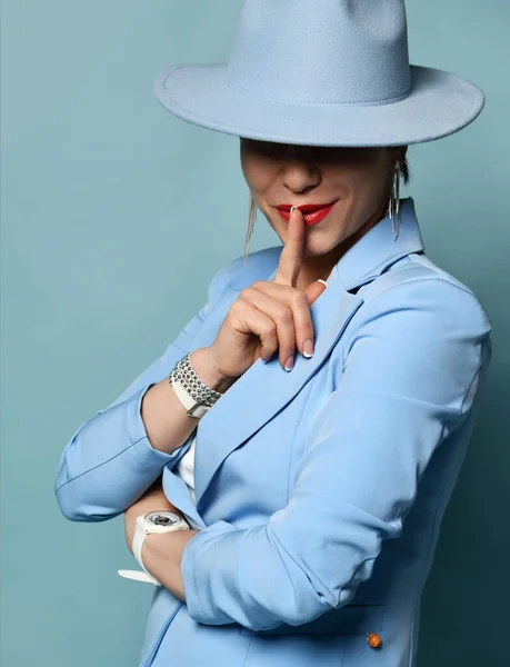 Slyly sorrindo mulher morena de cabelos curtos em azul negócio terno casual inteligente e chapéu de pé gesto sinal de silêncio — Fotografia de Stock