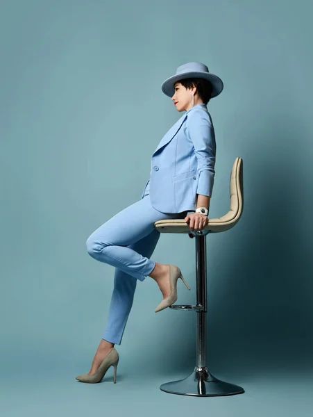 Rövid hajú barna nő kék öltönyben, kalap, magas sarkú cipő ül a bárszéken. Teljes növekedés. Oldalnézet — Stock Fotó