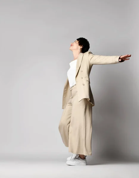 Happy adult woman in beige business casual pantsuit and sneakers stands sideways, dancing. Stylish business female wear —  Fotos de Stock