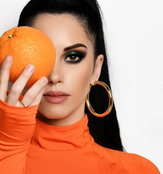 Молода брюнетка в сучасному помаранчевому светрі, сорочці і сережках тримає апельсин перед її обличчям — стокове фото