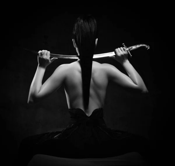 Черно-белый портрет молодой красивой брюнетки, сидящей топлесс к камере с мечом на плечах — стоковое фото