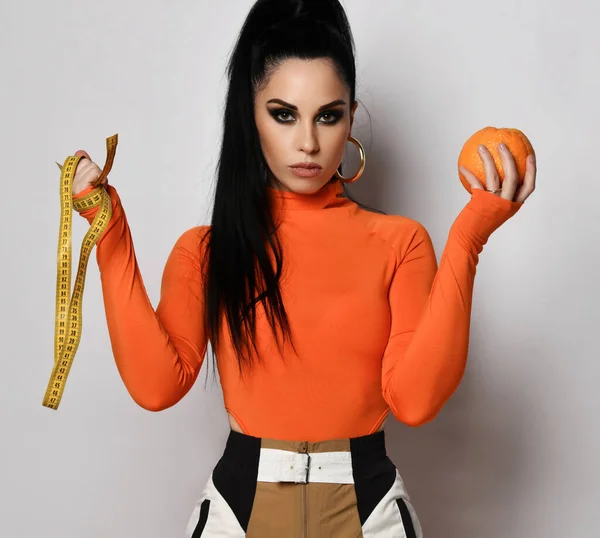 Mladá štíhlá žena v jasně oranžové sportovní oblečení kalhoty a rolák stojí drží čerstvou oranžovou a měřicí pásku v rukou — Stock fotografie
