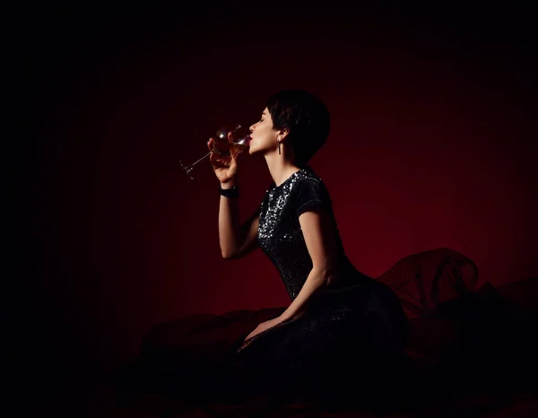 Adult pretty short haired brunette woman in black tulle dress sits on knees on floor drinking white wine. Side view — Foto de Stock