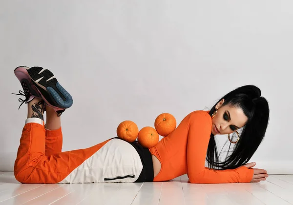 Jovem magro morena de cabelos longos em sportswear laranja e tênis está deitado no chão com laranjas frescas nas costas — Fotografia de Stock