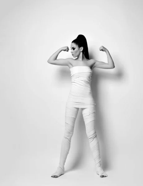 Young brunette woman in white medical bandages on body, body wrap stands showing her biceps. Black and white