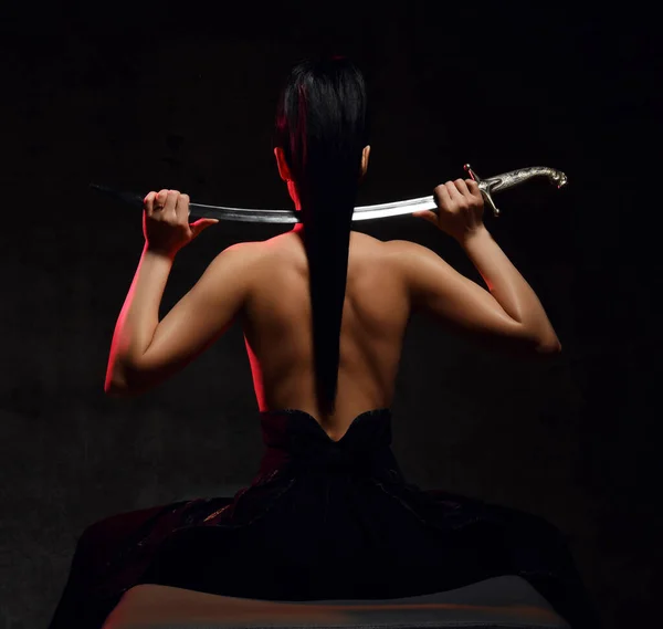 Vista posterior de la joven guapa morena guerrera sentada en topless de nuevo a la cámara sosteniendo la espada sobre los hombros —  Fotos de Stock
